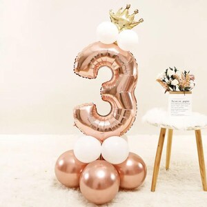 May include: A rose gold number three balloon with a gold crown on top. The balloon is surrounded by white and rose gold balloons.