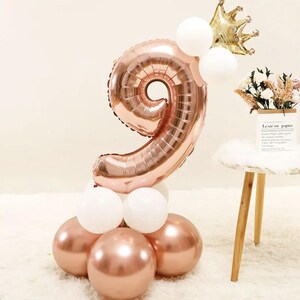 May include: A rose gold number 9 balloon display with white and rose gold balloons. A gold crown and flowers in a paper bag complete the festive arrangement. Ideal for a birthday celebration.