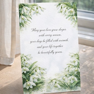 May include: A greeting card with a watercolor floral design featuring white snowdrop flowers and green leaves. The card has the text: "May your love grow deeper with every season, your days be filled with warmth, and your life together be beautifully yours."