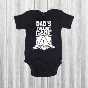 May include: Black baby bodysuit featuring the text "DAD'S PULLOUT GAME" in white, above a die graphic with the number 1 and the words "CRITICAL FAILURE" on a banner. The garment is set against a light wood backdrop.