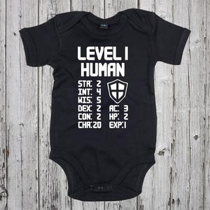 May include: A black baby growsuit with white text that reads "Level I Human" and includes a character sheet with stats like "STR: 2", "INT: 4", "WIS: 5", "DEX: 2", "CON: 2", "CHA: 20", "AC: 3", "HP: 2", and "EXP: 1".