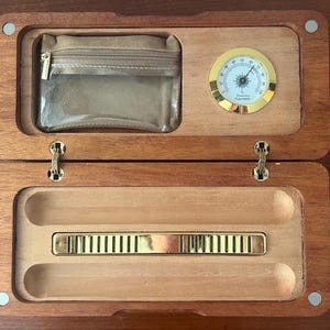 May include: Open wooden humidor with a clear pouch, a gold-rimmed hygrometer, and a brass vent. The humidor is open, revealing the interior with a brass vent and two curved grooves. The wood has a rich, warm tone.