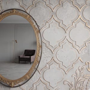 May include: An oval-shaped mirror with a gold ornate frame and a dark metal outer frame. The mirror reflects a room with a chair, a floor lamp, and a light gray wall. The wall to the right has a textured, light gray pattern with gold accents.