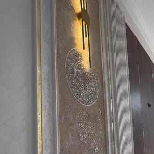 Puede incluir: Una pieza decorativa de arte mural con un diseño abstracto dorado y un patrón circular ornamentado. La pared tiene un acabado beige texturizado con elementos decorativos y está enmarcada por molduras blancas.