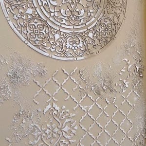May include: A textured beige wall featuring raised white patterns. The design includes a large circular motif at the top and a repeating geometric pattern below, creating an artistic surface. The patterns are raised.