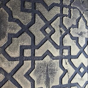 May include: Close-up of a textured surface with a repeating geometric pattern. The design features gold-colored shapes outlined by dark gray lines. The shapes include squares, diamonds, and star-like forms, creating a visually complex and detailed pattern.