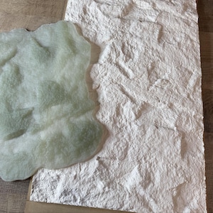 May include: Two textured surfaces are displayed. One is a large, off-white rectangular piece with a rough, stone-like appearance. The other is a smaller, irregularly shaped piece in a pale green color, with a soft, plush texture. Both are on a light brown surface.