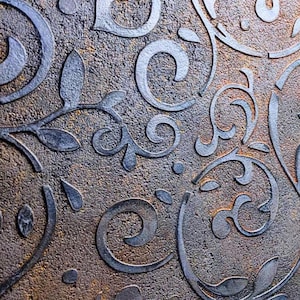 Può includere: Primo piano di una superficie strutturata con un motivo ripetuto di disegni floreali e a volute stilizzati. Gli elementi in rilievo sono grigio metallizzato, in contrasto con lo sfondo marrone e oro screziato. Il design ha un'estetica vintage o antica.