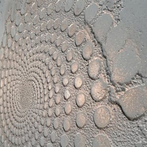May include: Close-up of a textured, silver-colored wall with a spiral pattern of raised, oval-shaped elements. The design creates a sense of depth and movement, with the elements diminishing in size towards the center.