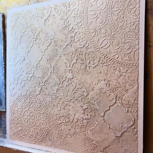 May include: A square, textured wall art piece in beige and off-white. The artwork features a raised, intricate pattern of floral and geometric designs, including circular and quatrefoil shapes, creating a tactile and decorative surface.