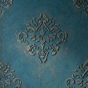 May include: A deep teal wall panel featuring a raised, textured damask pattern in a distressed gold finish. The surface has a rough, hand-applied plaster texture, with the ornate floral motifs centered and repeated in the corners of the panel.