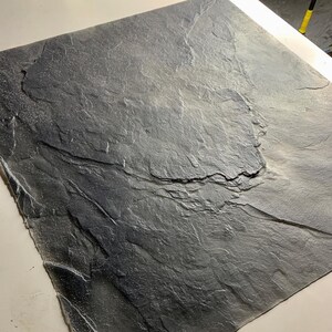 May include: A square, dark gray tile featuring a textured, uneven surface that mimics the appearance of natural slate stone. The material displays a matte finish with subtle variations in tone and deep, jagged ridges across the entire face.