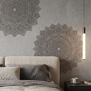 May include: Two large, intricate mandala wall decals in dark gray adorn a light gray textured wall. The decals are above a bed with neutral-toned bedding. A modern black nightstand with a lamp and a cup of coffee is visible.