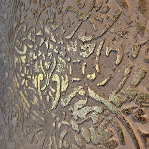 Puede incluir: Primer plano de una pared texturizada con una base marrón y detalles de pan de oro. El diseño presenta un patrón ornamental y repetitivo de elementos florales y de volutas, creando una estética vintage o antigua. El pan de oro añade un toque de lujo.