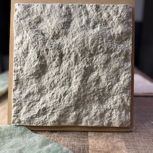 May include: A square, light beige stone tile with a rough, textured surface. The tile is set within a brown cardboard frame, showcasing the natural stone's rugged appearance. The tile is likely for home decor or construction.