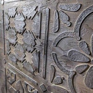 May include: Close-up of a textured, metallic surface with an intricate, carved design. The design features geometric shapes, starburst patterns, and floral elements. The color palette includes shades of gray, brown, and hints of gold, creating a rustic, aged appearance.