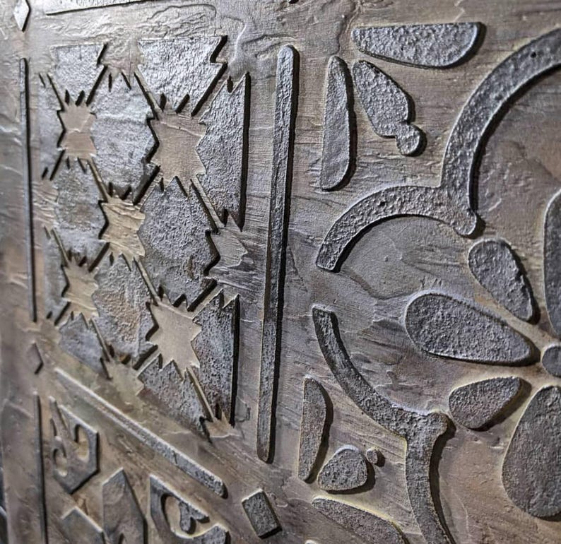 May include: Close-up of a textured, metallic surface with an intricate, carved design. The design features geometric shapes, starburst patterns, and floral elements. The color palette includes shades of gray, brown, and hints of gold, creating a rustic, aged appearance.