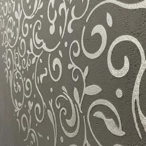 Puede incluir: Primer plano de una pared gris con un patrón floral plateado repetido. El diseño presenta líneas onduladas, hojas y formas circulares, creando una superficie decorativa texturizada. El patrón se aplica a la superficie de la pared.
