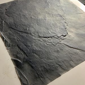 Op de afbeelding: Een vierkante, getextureerde, donkergrijze leistenen tegel. Het oppervlak heeft een natuurlijk, gelaagd uiterlijk met variaties in toon en textuur. De tegel is waarschijnlijk bedoeld voor gebruik in de bouw of voor interieurdecoratie.
