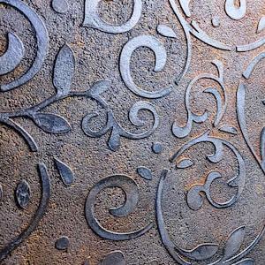 Może przedstawiać: Zbliżenie teksturowanej powierzchni z powtarzającym się wzorem stylizowanych kwiatowych i przewijanych wzorów. Podniesione elementy mają metaliczny srebrny kolor, kontrastujący z brązowym i złotym tłem. Projekt ma estetykę vintage.