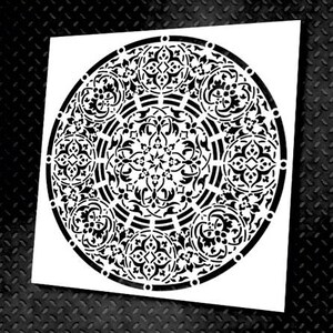 May include: A white stencil featuring a detailed black circular design. The design is an intricate pattern of floral and geometric shapes, creating a mandala-like appearance. The stencil is on a black textured surface.