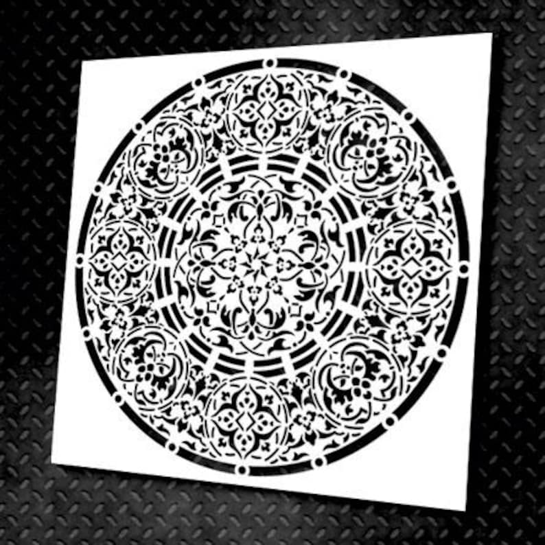 May include: A white stencil featuring a detailed black circular design. The design is an intricate pattern of floral and geometric shapes, creating a mandala-like appearance. The stencil is on a black textured surface.