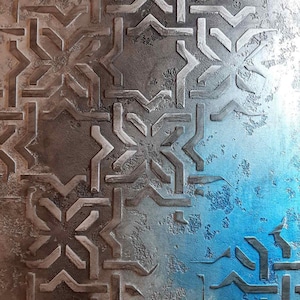 Può includere: Superficie metallica strutturata con un motivo geometrico ripetuto. Il design presenta forme angolari e linee in colore grigio metallizzato, con una sfumatura blu sul lato destro.
