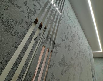 May include: A modern interior wall design featuring vertical, reflective metal strips against a textured gray wall. The strips are arranged in a parallel pattern, creating a sense of depth and dimension. A recessed light fixture is visible.