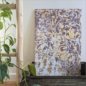 May include: A rectangular canvas art piece with a distressed design. The artwork features a pattern of gold floral motifs on a deep purple background, with white and gold accents. The piece is displayed in a rustic wooden box.