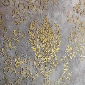 May include: Close-up of a textured wall with a gray background and gold floral patterns. The design features a central floral arrangement and ornate, swirling leaf and vine motifs, creating a classic, elegant aesthetic.
