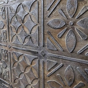 May include: Close-up of a wooden surface with a repeating pattern of geometric shapes. The design features diamond, oval, and linear elements in a raised, textured relief. The color palette is a mix of brown and gray, creating a rustic, aged appearance.