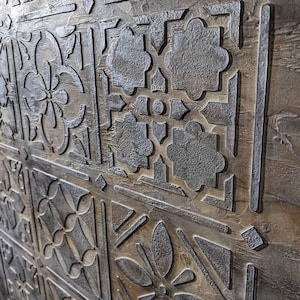 May include: Close-up of a decorative wooden panel with a repeating pattern of carved floral and geometric designs. The panel has a weathered, textured appearance with shades of brown and gray, creating a rustic aesthetic.