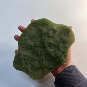 Puede incluir: Una piedra verde de forma irregular con una superficie texturizada. La piedra se sostiene en una mano, mostrando su tamaño y forma. El color es un verde natural apagado, lo que sugiere jade o un material similar. Los bordes de la piedra son redondeados.