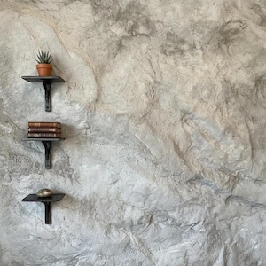 Puede incluir: Tres pequeñas estanterías de metal negro están montadas en una pared de piedra gris texturizada. La estantería superior contiene una pequeña suculenta en maceta. La estantería del medio tiene una pila de libros. La estantería inferior muestra una piedra lisa y redonda.