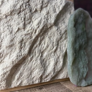 May include: Close-up of two textured stone pieces. One is a large, light beige rectangular block with a rough, uneven surface. The other is a smaller, rounded, pale green stone. The stones are set against a wooden surface.