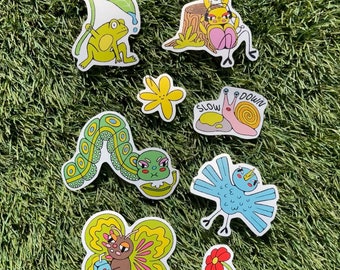 May include: A set of eight colorful stickers featuring various animals and plants. The stickers include a frog under a leaf, a snail with the words "slow down", a caterpillar, a butterfly, a bird, a flower, and a deer on a tree stump.