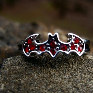 May include: A silver ring with a red rhinestone bat design.
