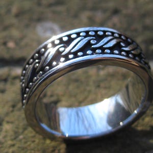 May include: A silver-colored ring with an ornate design. The ring features a band with a repeating pattern of dots and leaf-like shapes. The ring is highly polished and reflects the surrounding environment.