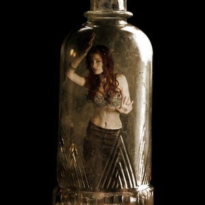 Puede incluir: Una botella de vidrio transparente con tapón de corcho, con una mujer dentro. La mujer tiene el pelo largo y rojo y lleva un top y una falda decorativos. La botella tiene una base texturizada y diseños geométricos.