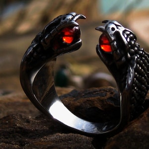 May include: A silver ring with two cobra heads, each with a red gemstone. The snake heads face each other. The ring is set against a blurred background, emphasising the detailed design of the snake heads.