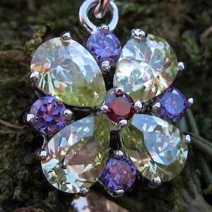 May include: A silver pendant necklace with a floral design. The pendant features pear-shaped, light green gemstones, purple gemstones, and a central red gemstone. The necklace is set against a mossy background.