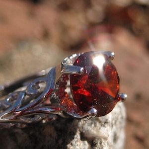 May include: Close-up of a silver ring with a large, oval-cut, deep red gemstone. The ring has an ornate, filigree design on the band. The gemstone is the focal point, catching the light and reflecting a fiery glow.