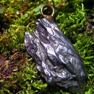 Könnte beinhalten: Dunkler, strukturierter Metallanhänger mit Schlaufe, eingebettet in grünes Moos. Der Anhänger hat eine raue, organische Form mit metallischem Glanz, was auf ein natürliches oder handgefertigtes Design hindeutet.