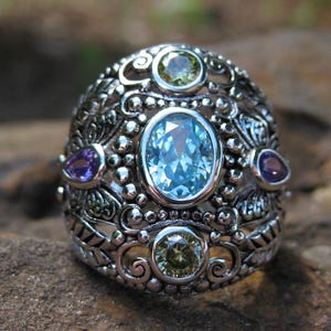 Può includere: Un anello d'argento con un designo dettagliato che presenta cinque pietre preziose. L'anello ha una grande pietra preziosa blu al centro, circondata da pietre preziose verdi, viola e verdi più piccole.