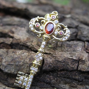 May include: A gold-colored key with an ornate design, featuring a central oval red gemstone and numerous smaller clear gemstones. The key's handle and head are embellished with intricate details, creating a vintage or antique aesthetic.