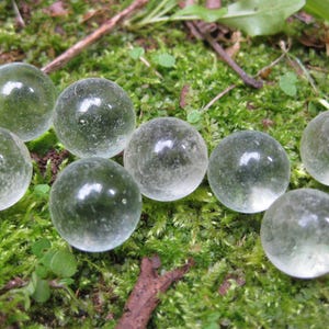 Puede incluir: Ocho canicas de vidrio transparente con un ligero tinte verde están dispersas sobre un lecho de musgo verde. Las canicas tienen una superficie lisa y reflectante, y algunas tienen inclusiones internas.