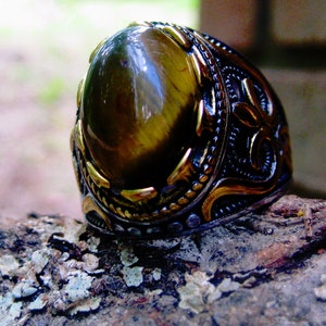May include: A close-up of a tiger's eye gemstone ring. The ring features a large, oval-shaped, polished tiger's eye stone set in a dark metal band with gold-colored filigree and detailing. The ring is resting on a textured, natural surface.