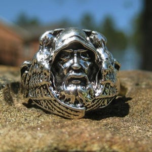 May include: A silver-colored ring featuring a detailed depiction of a hooded figure with a long beard. The ring has an intricate design with a weathered appearance, set against a natural, textured background. The ring's design is centered on the face of the figure.