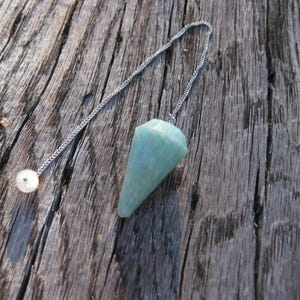 May include: A faceted, light green, cone-shaped gemstone pendulum attached to a thin silver-colored metal chain. The chain features a small, clear, spherical bead at the end, providing a balanced weight for the hanging stone pendant.