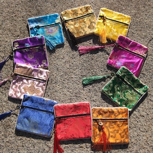 Puede incluir: Una colección de pequeñas bolsas cuadradas de satén en varios colores, incluyendo azul, dorado, amarillo, rosa, verde y rojo. Cada bolsa tiene una cremallera negra y una borla a juego. Las bolsas están dispuestas en un patrón circular.